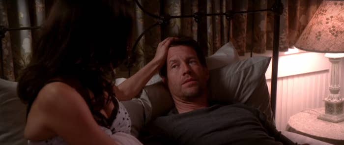 A woman in a polka dot dress sits on a bed beside a man lying down, looking up at her, in a dimly lit room