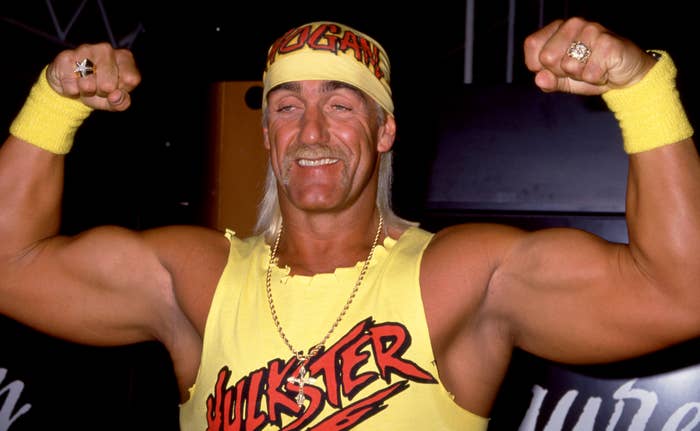 Person flexing in a sleeveless shirt and bandana, both with bold logos, smiling with confident posture in a casual setting