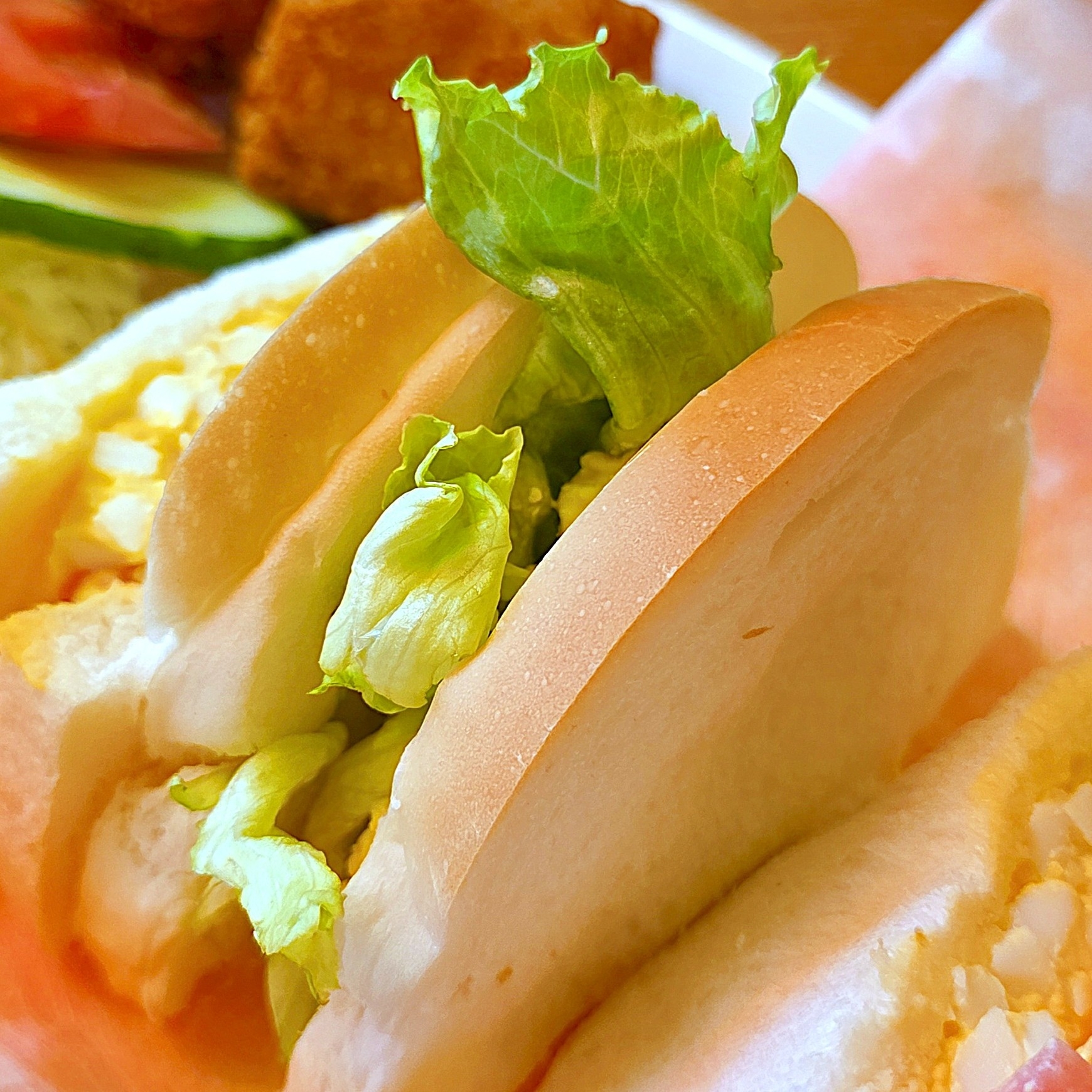 サンドイッチにレタスと卵サラダが挟まれた至近距離の写真。背景に野菜と揚げ物が見える。
