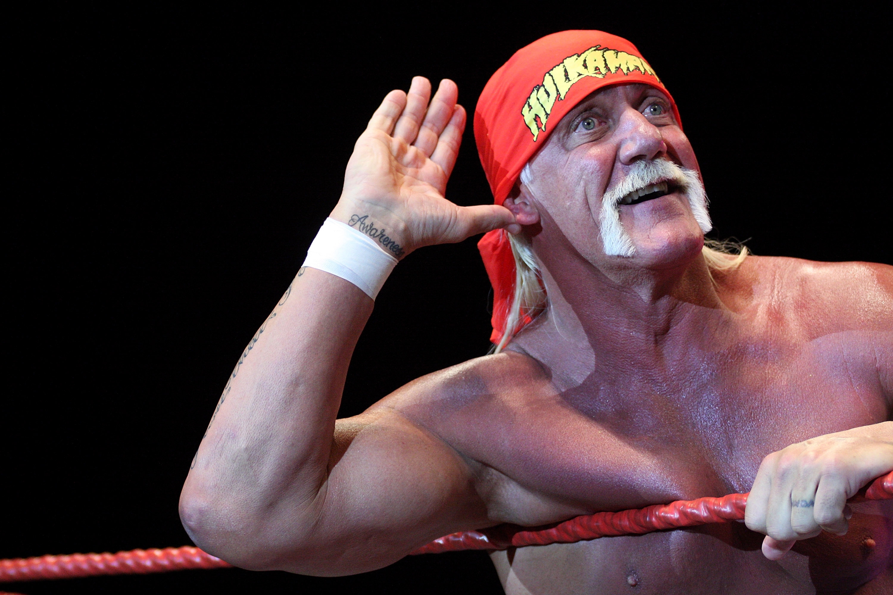 A wrestler with a handlebar mustache and red bandana cups his ear while leaning on a wrestling ring rope