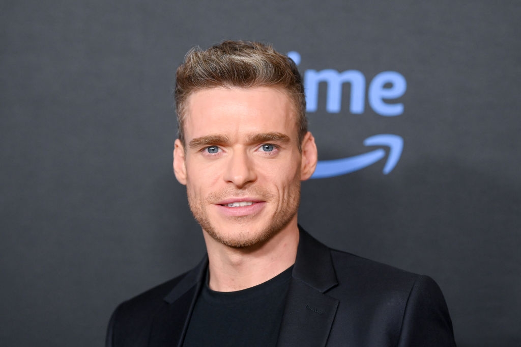 Man in a black suit with a black shirt at a media event, standing in front of a backdrop with the Amazon Prime logo
