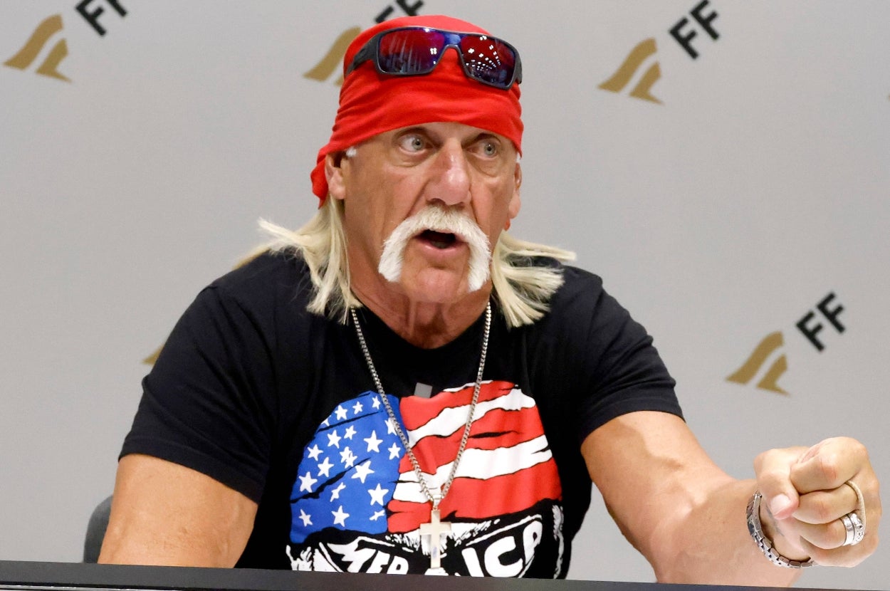 Person wearing a bandana and sunglasses on head, with a mustache and graphic t-shirt, gesturing during an event or interview