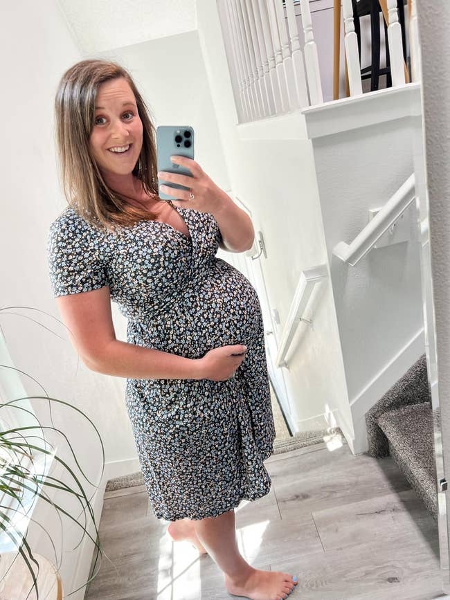 Person taking a mirror selfie, holding their baby bump, wearing a floral dress, barefoot indoors near stairs