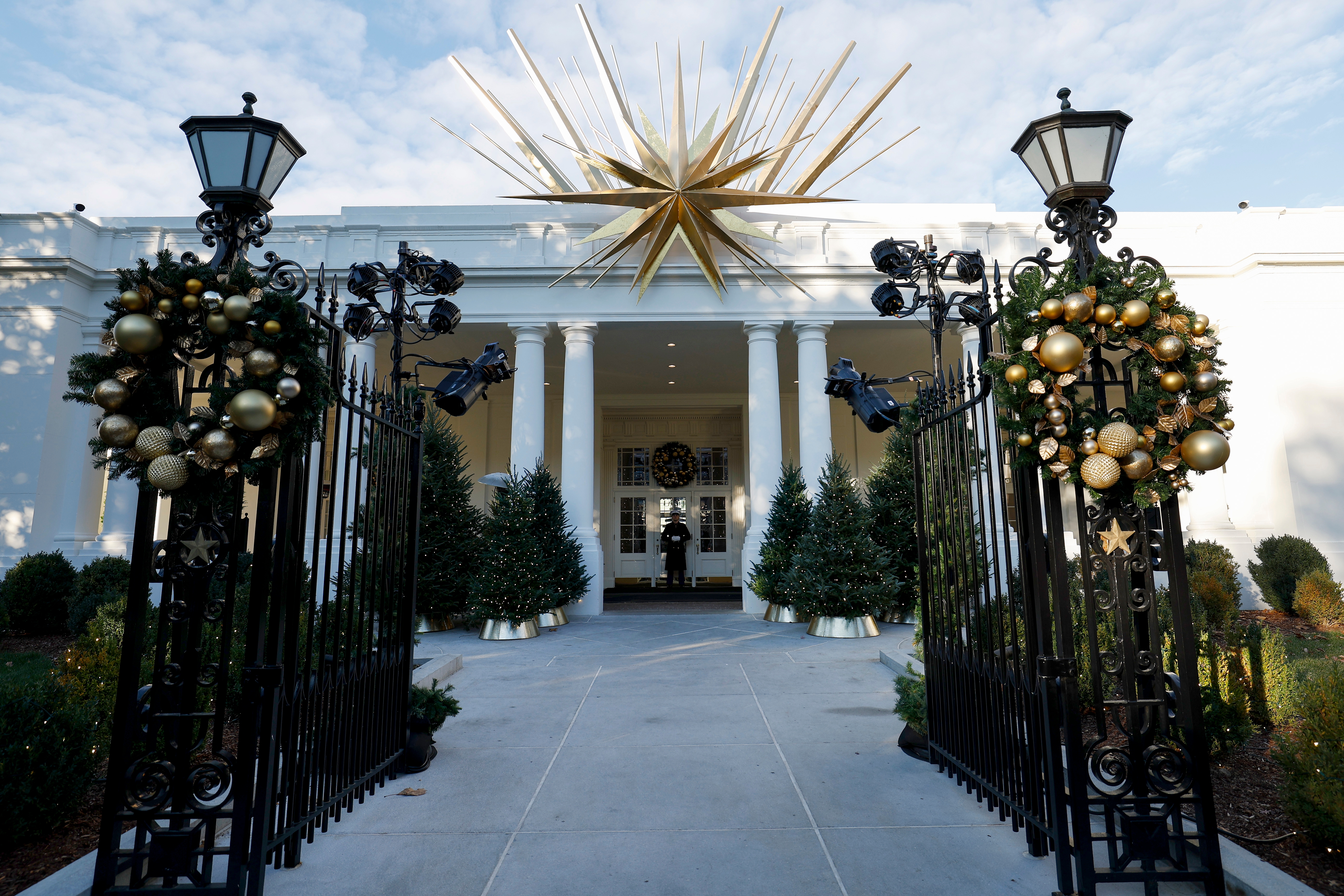 White House entrance decorated with stars, garlands, and a large golden ornament above, seen through ornate open gates