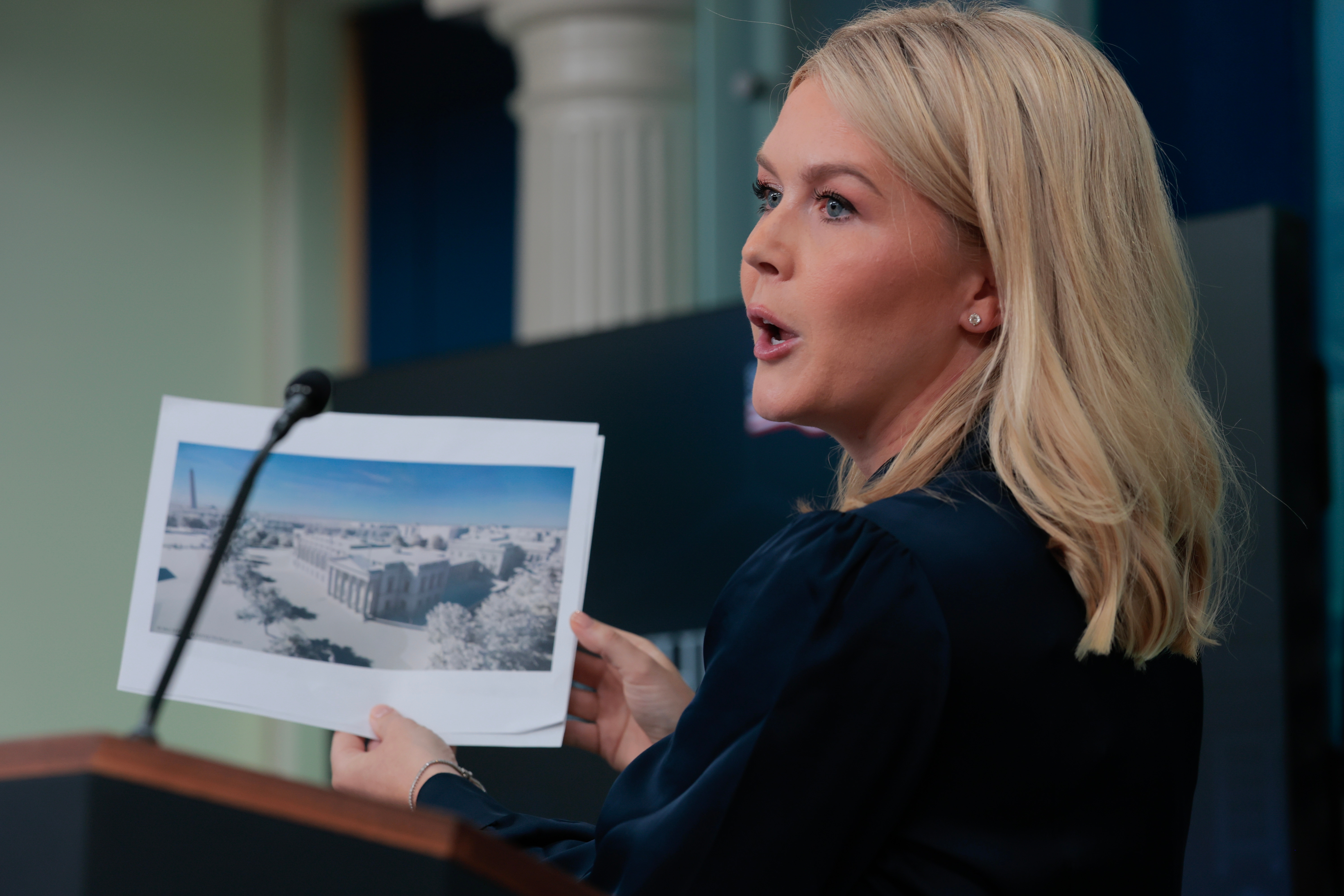 Person speaking at a podium holds a photo of a snowy landscape in a government or press briefing setting
