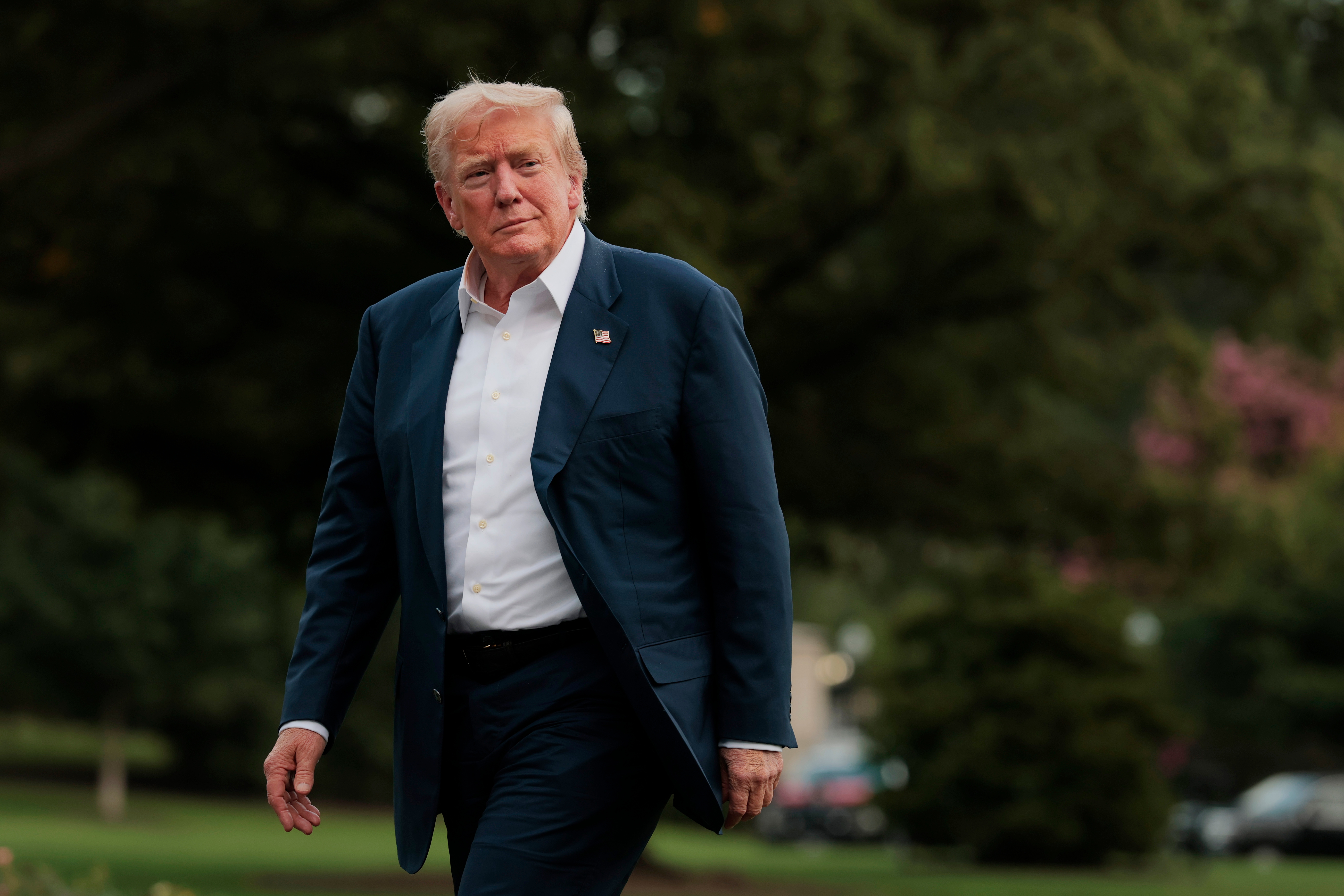 A person walking outside in a suit, looking ahead. Trees and blurred greenery in the background