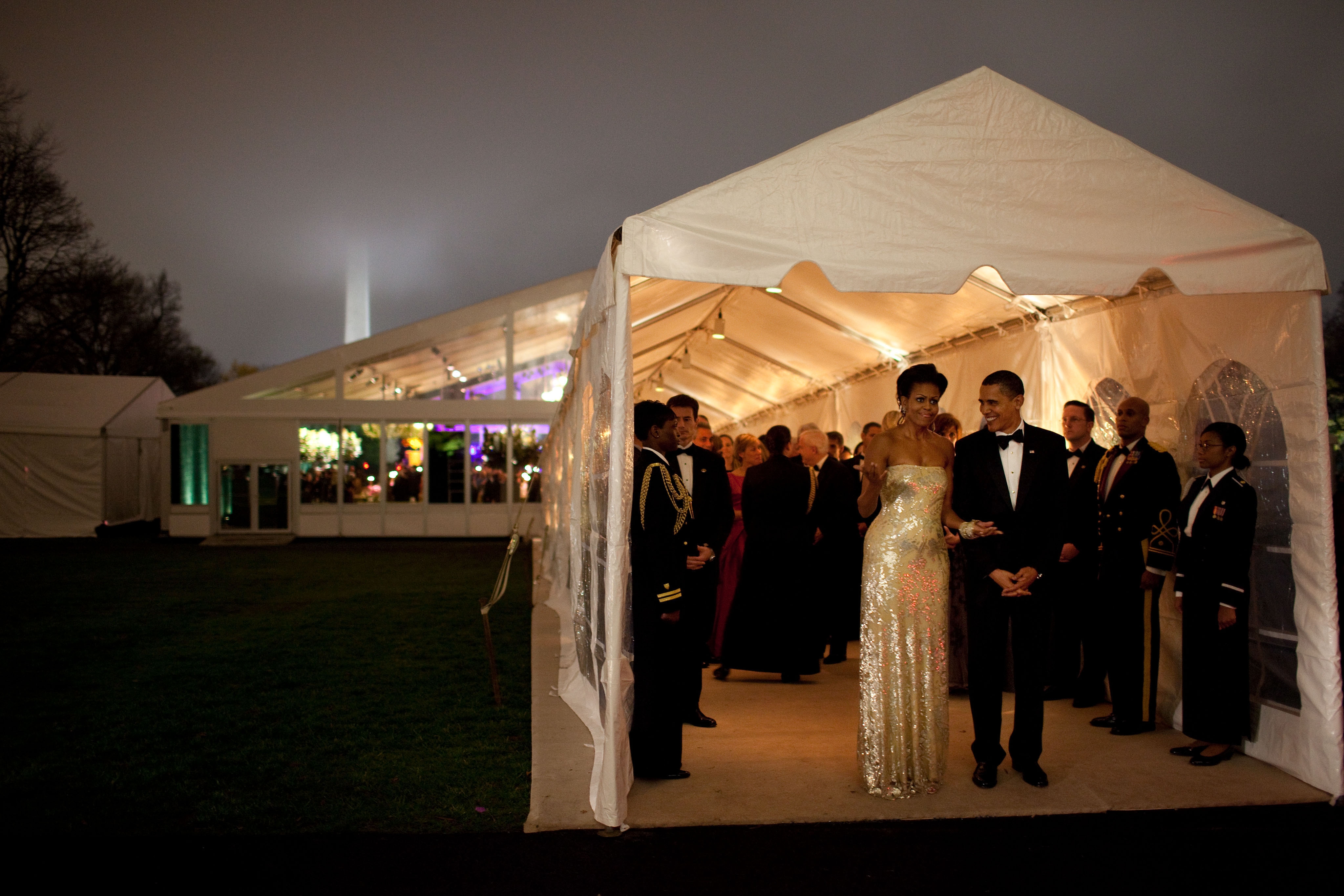 People in formal wear entering a lit marquee at an event, with a tall building visible in the background