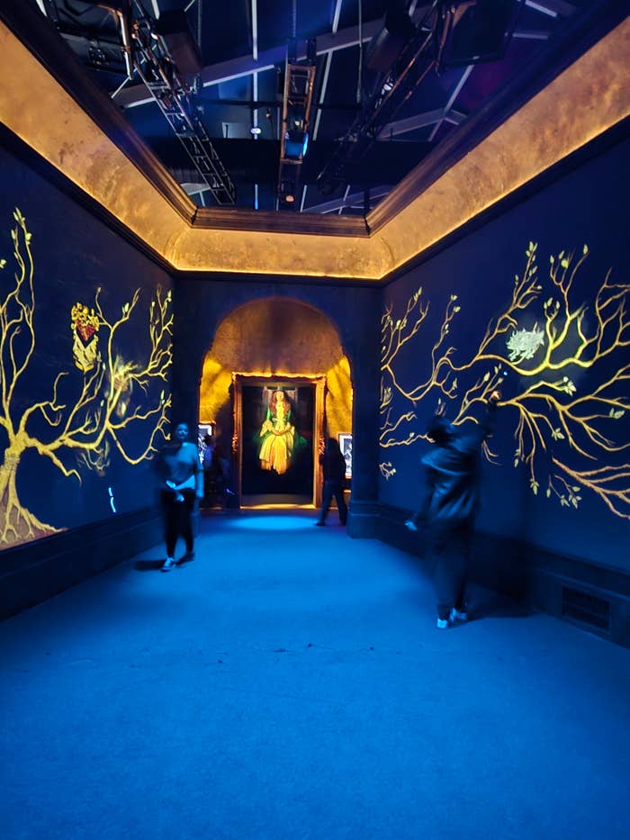 Galería con decoración de ramas pintadas en las paredes, personas observando una obra de arte iluminada al fondo