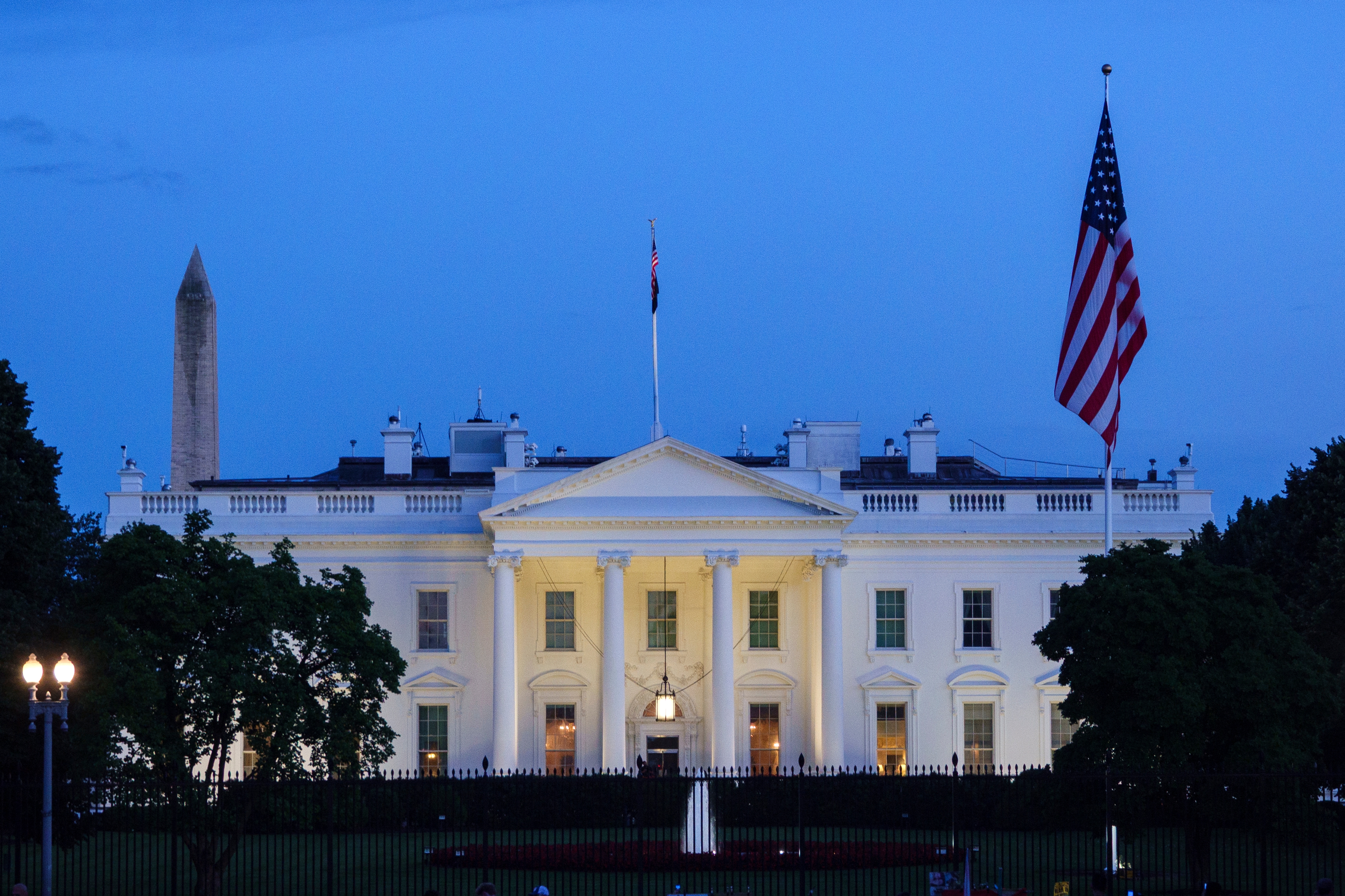 The White House at dusk with illuminated exterior, an American flag, and the Washington Monument in the background
