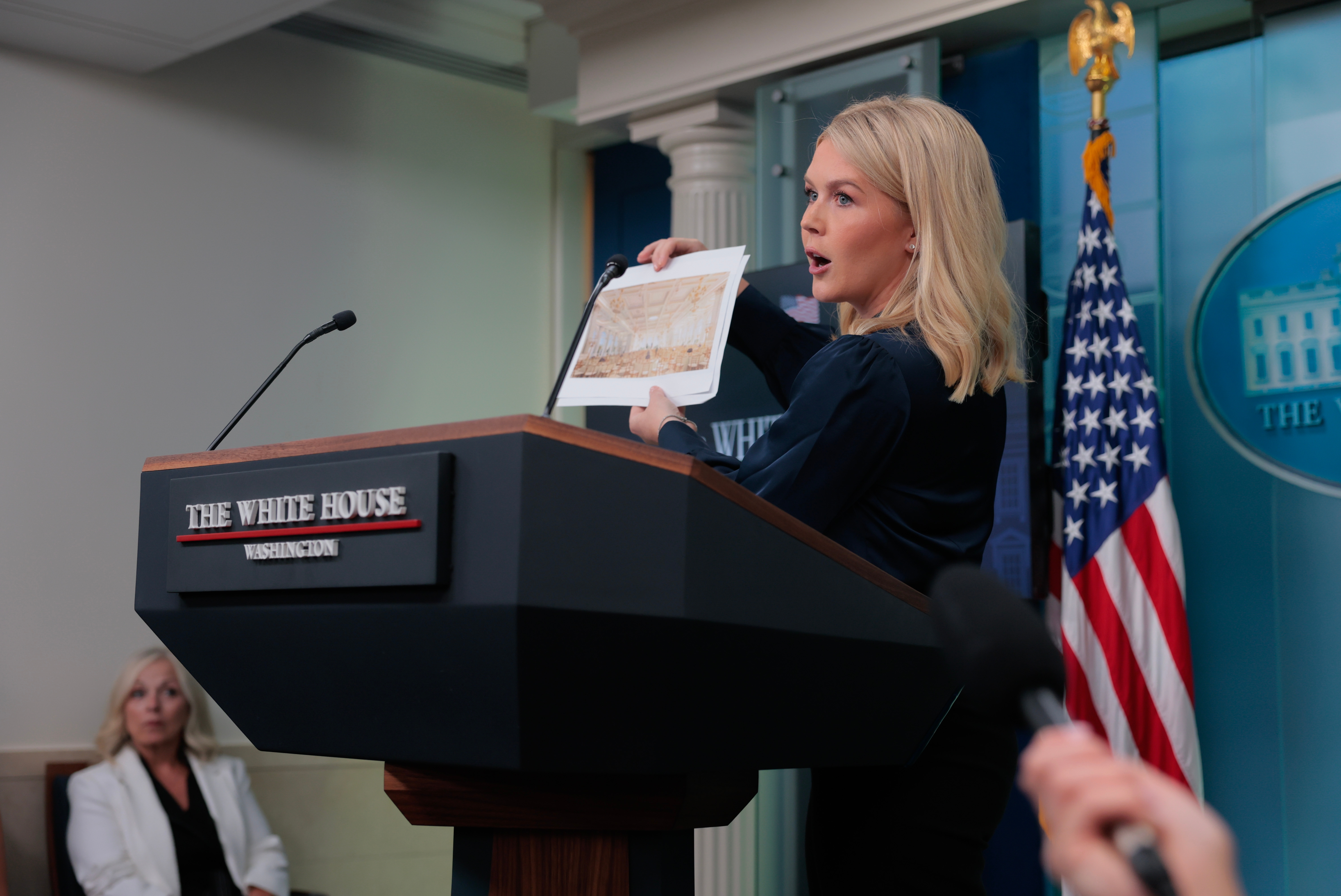 A woman at a podium holds a document during a White House press briefing, with a seated audience member and U.S. flag in the background