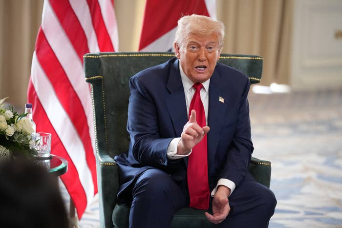 A person in a suit is seated, gesturing while speaking, with flags in the background