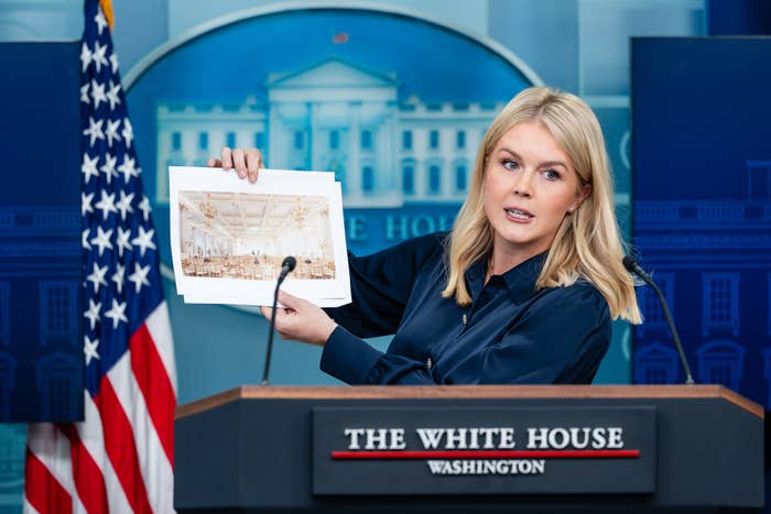 A person at a White House podium holds up an image of a room design. An American flag is visible on the side