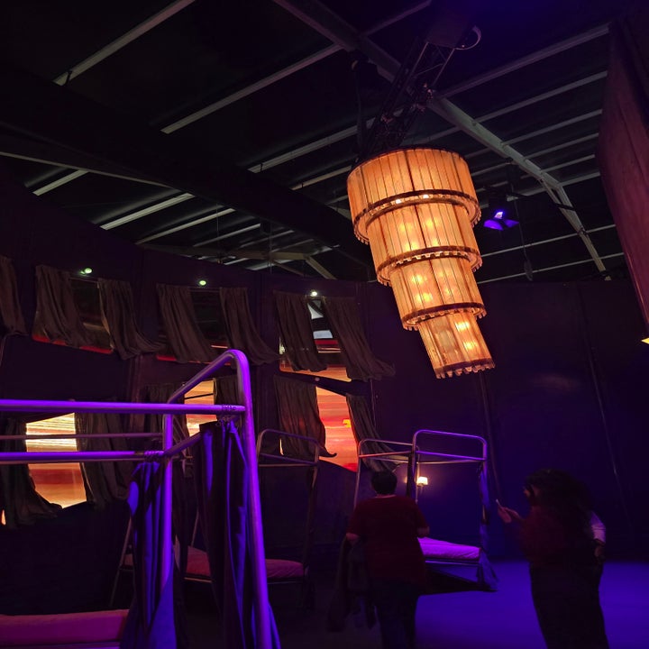 Personas caminando en una sala oscura con camas colgantes y una lámpara colgante grande. Ambiente teatral y moderno