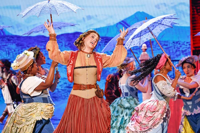 A performer in a vintage-inspired costume sings energetically on stage, surrounded by others with parasols in a choreographed scene