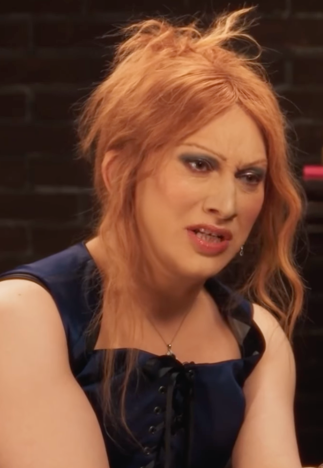 Person with long hair and expressive makeup, wearing a sleeveless top, appears to be speaking in an indoor setting
