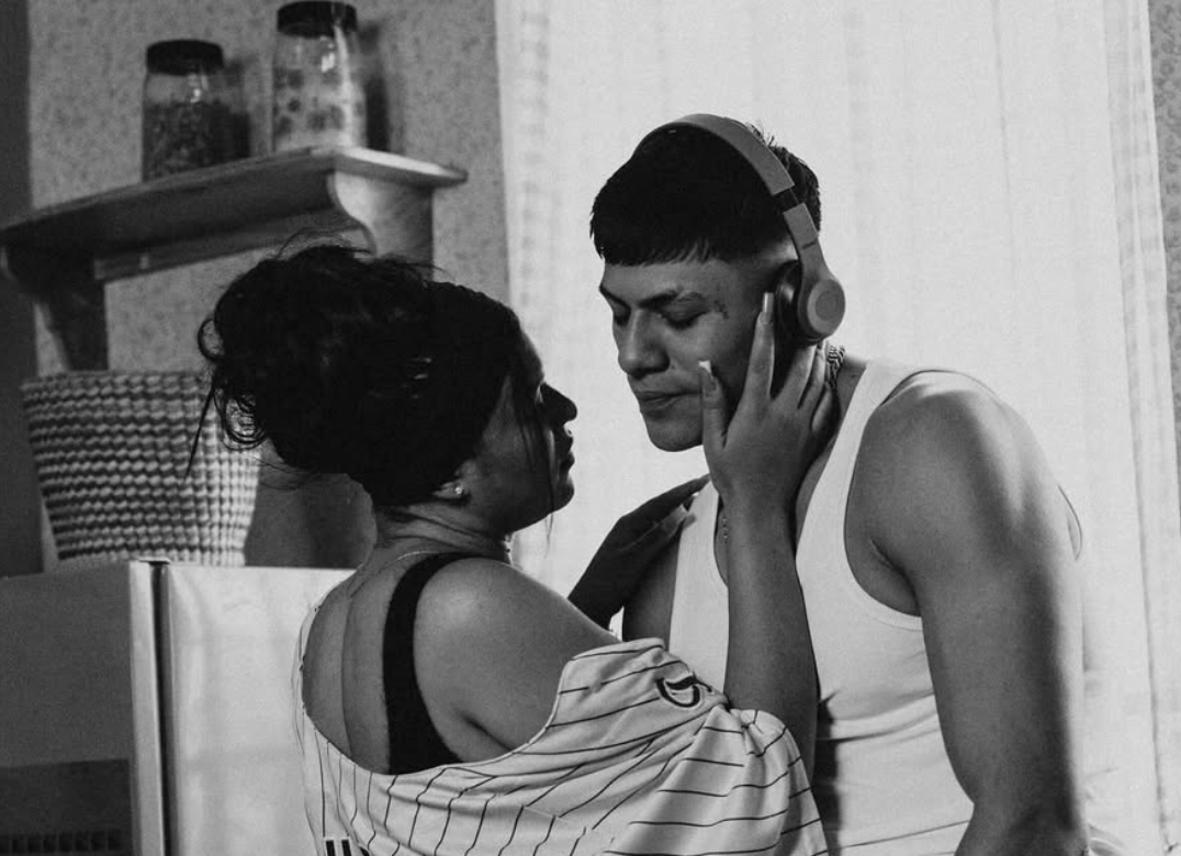 Pareja en cocina, mujer acaricia el rostro del hombre que usa audífonos, ambos tienen una expresión seria e íntima