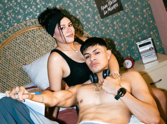 Persona con auriculares y reloj posando sin camisa junto a otra persona en vestido informal. Ambos parecen relajados sobre una cama