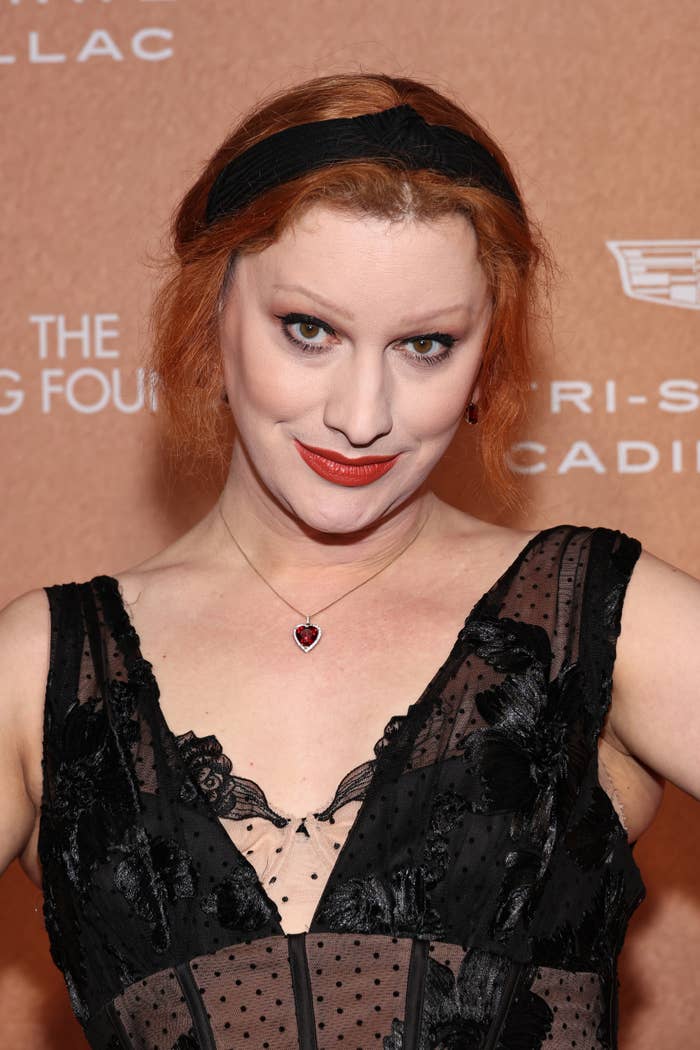 Person wearing a lacy dress with mesh panels and a heart necklace on a formal event backdrop