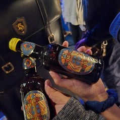 Dos manos sostienen botellas de Butterbeer, bebida famosa de Harry Potter, con etiquetas detalladas. Mochila en el fondo