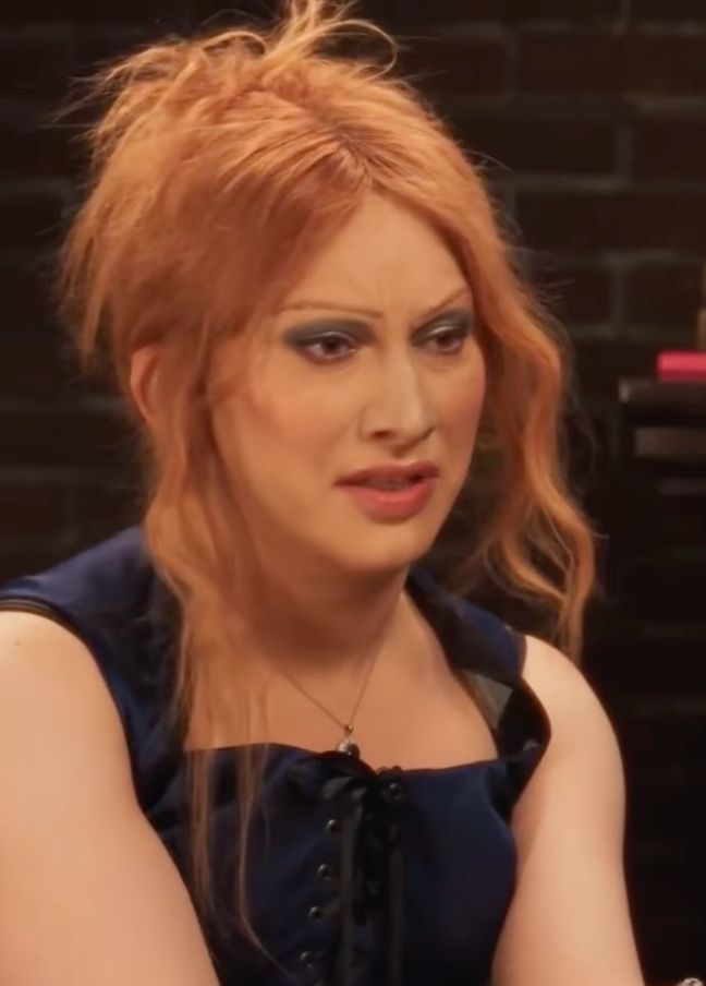 Person with long hair in a loose updo listening attentively, wearing a sleeveless dress with lace-up detail. Brick wall background