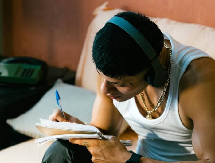 Persona con auriculares, escribiendo en un cuaderno mientras está sentado en un sofá, concentrado en su tarea. Lleva camiseta sin mangas y cadenas