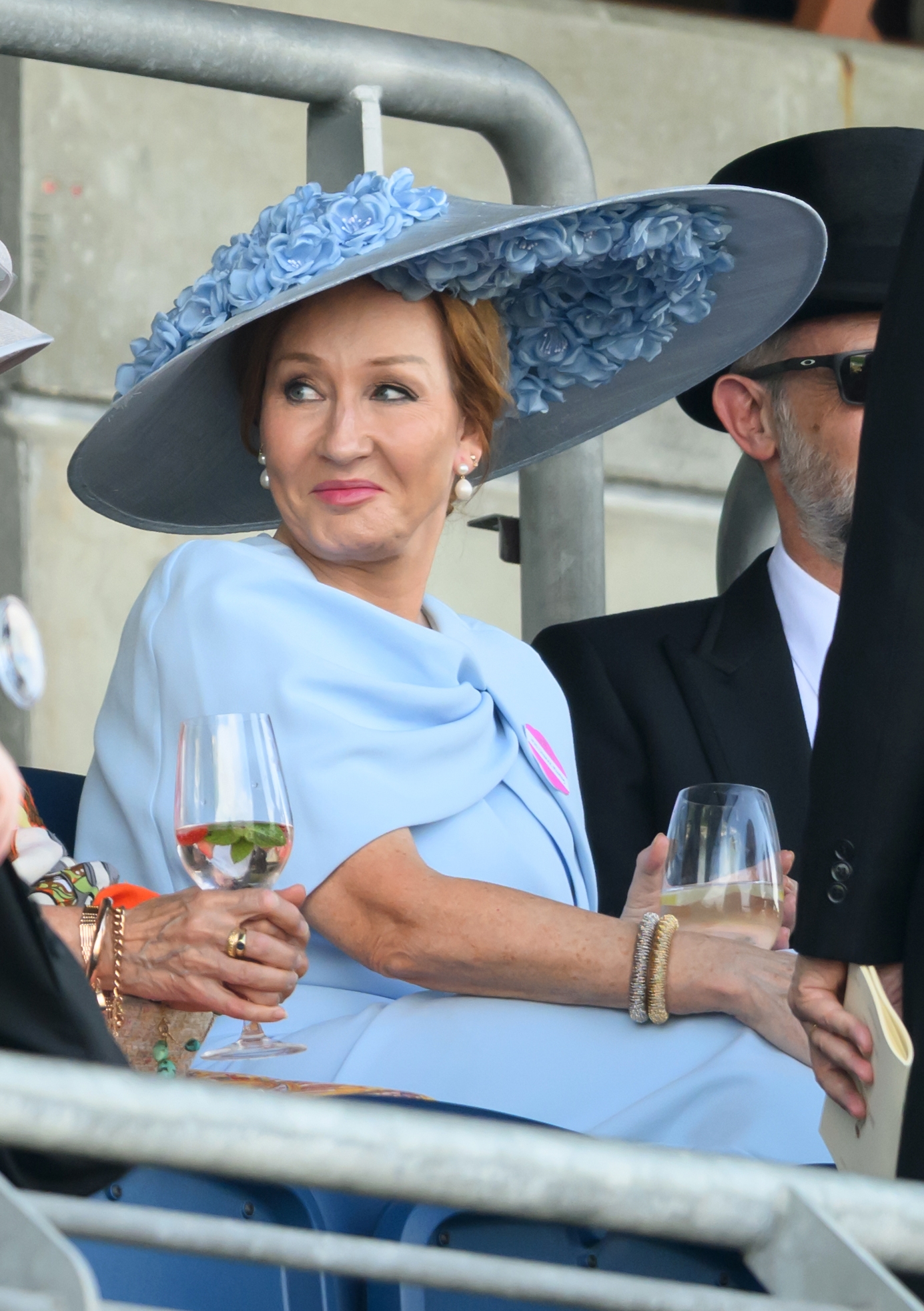 A woman in an elegant outfit with a wide-brimmed floral hat sits at an event, holding a drink, with others nearby