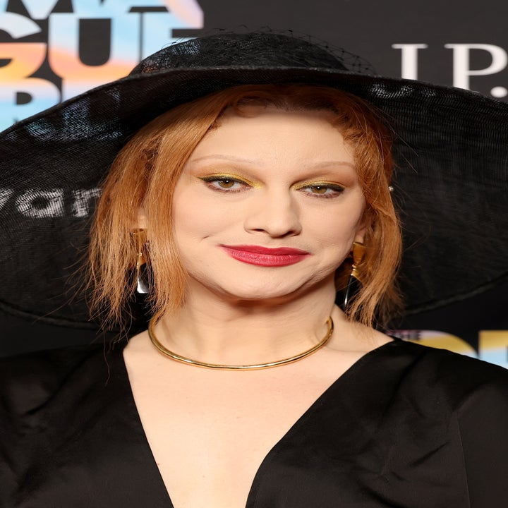 Person at Drama League Awards wearing a wide-brimmed black hat and elegant black outfit, standing in front of a branded backdrop
