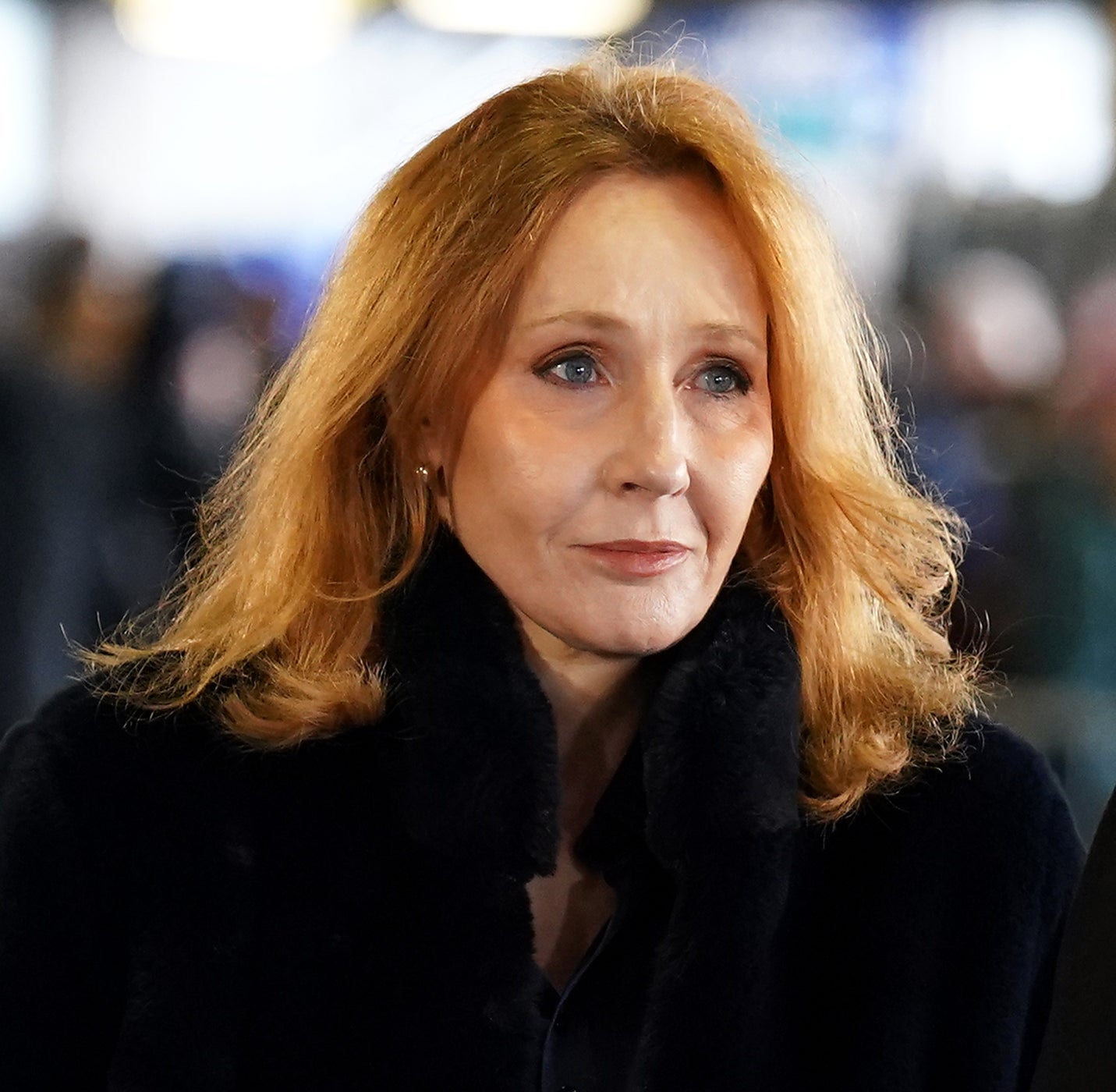 A person with long hair wearing a dark coat is standing in a crowded area, looking ahead