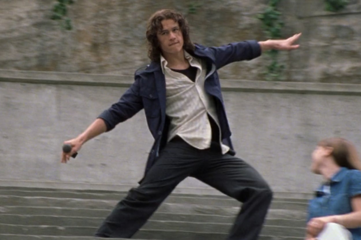 Person dancing energetically outdoors, wearing a dark jacket over a light shirt and holding a microphone