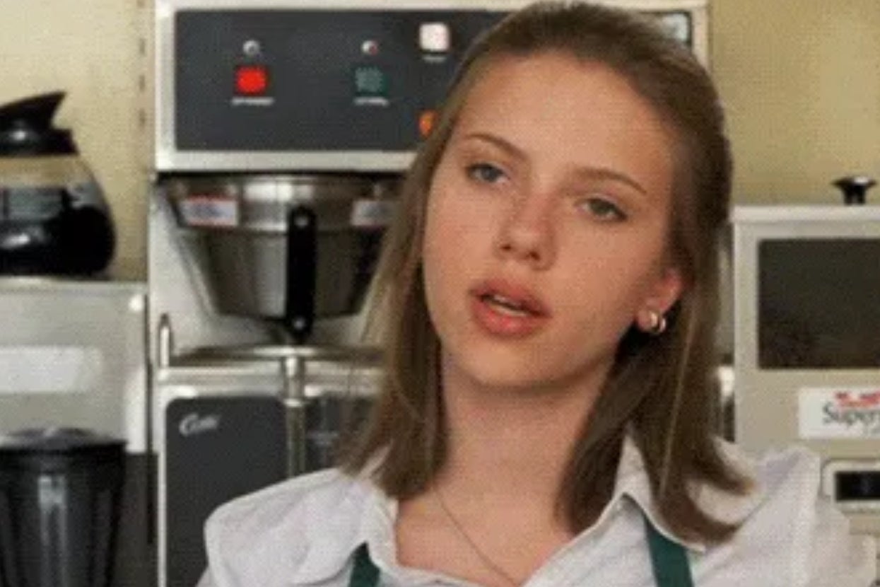 A person wearing a white shirt and green apron stands in a cafe setting, appearing focused on something