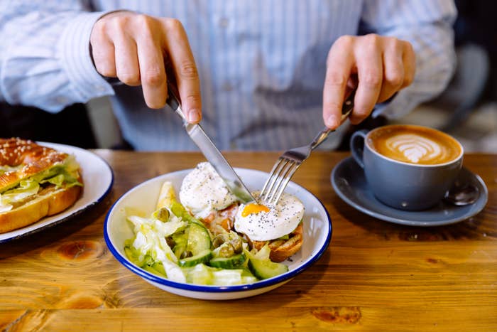 Person cutting into poached eggs on toast with avocado and salad; cappuccino and sandwich nearby on a wooden table