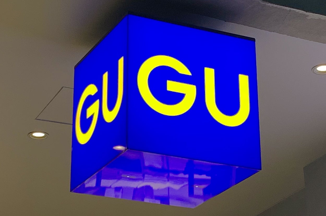 Cube-shaped sign with "GU" repeated in large yellow letters on a blue background, hanging indoors