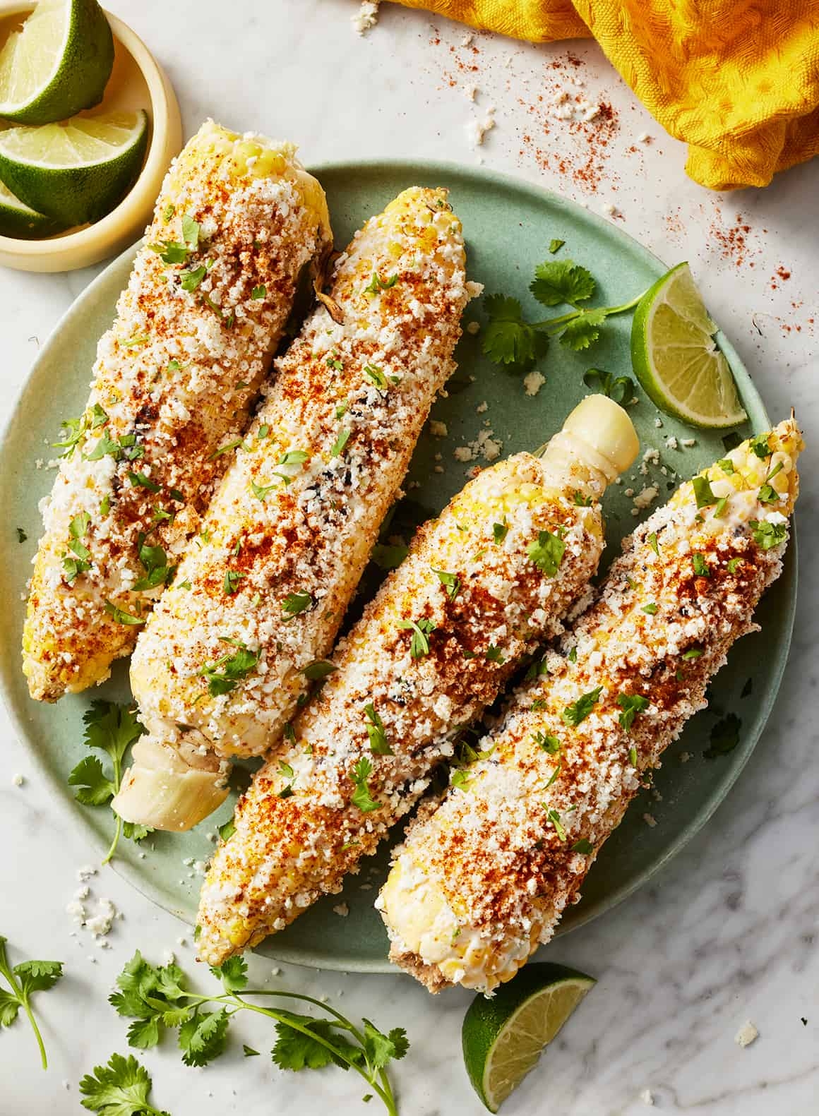 Four elotes (Mexican street corn) covered in cheese, chili powder, and cilantro on a plate with lime wedges