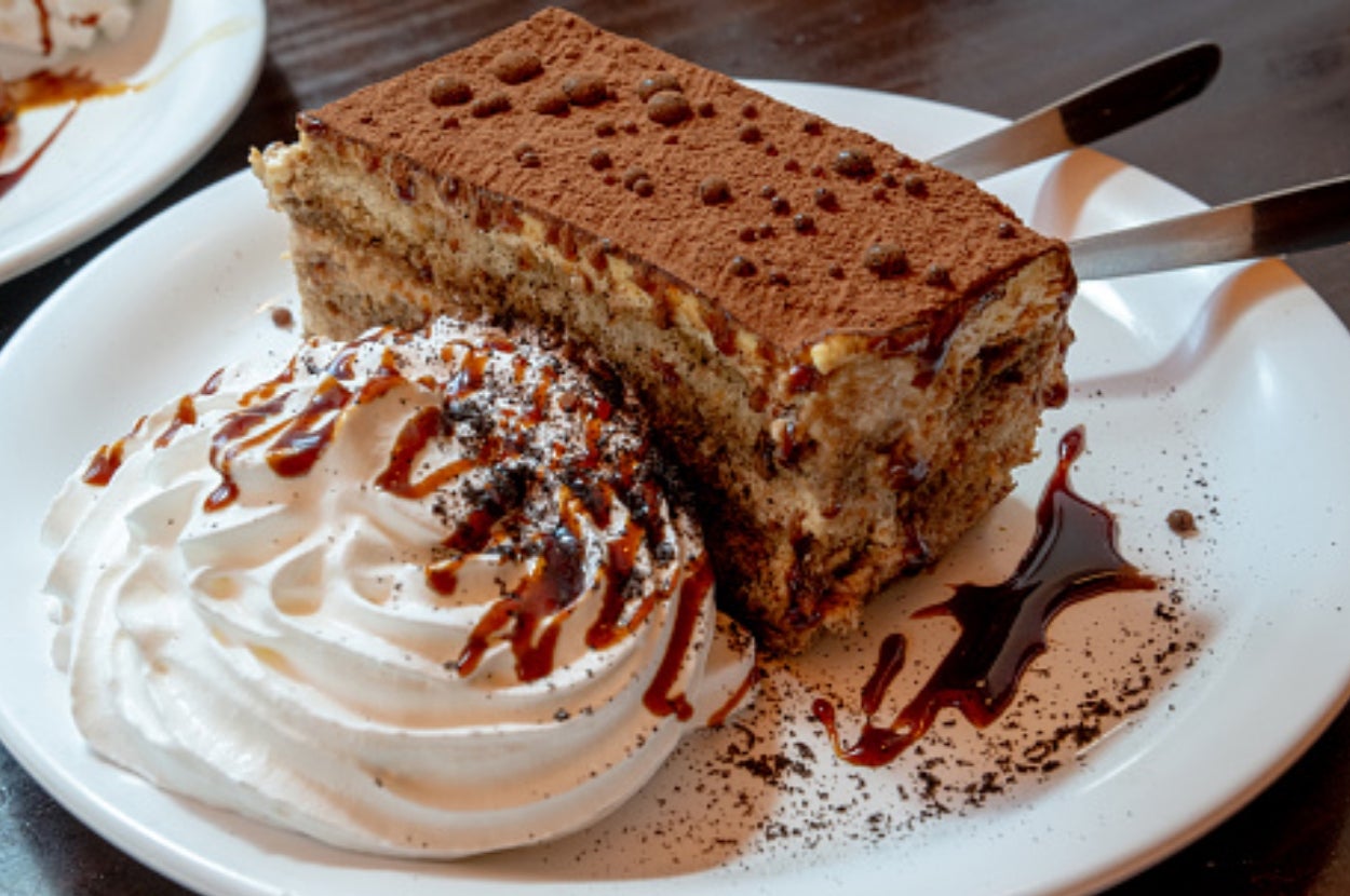 A slice of tiramisu on a plate with a swirl of whipped cream and chocolate sauce drizzle. Two spoons are placed beside the dessert