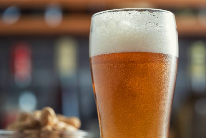 A frothy glass of beer sits on a bar counter, with blurred bar shelves in the background