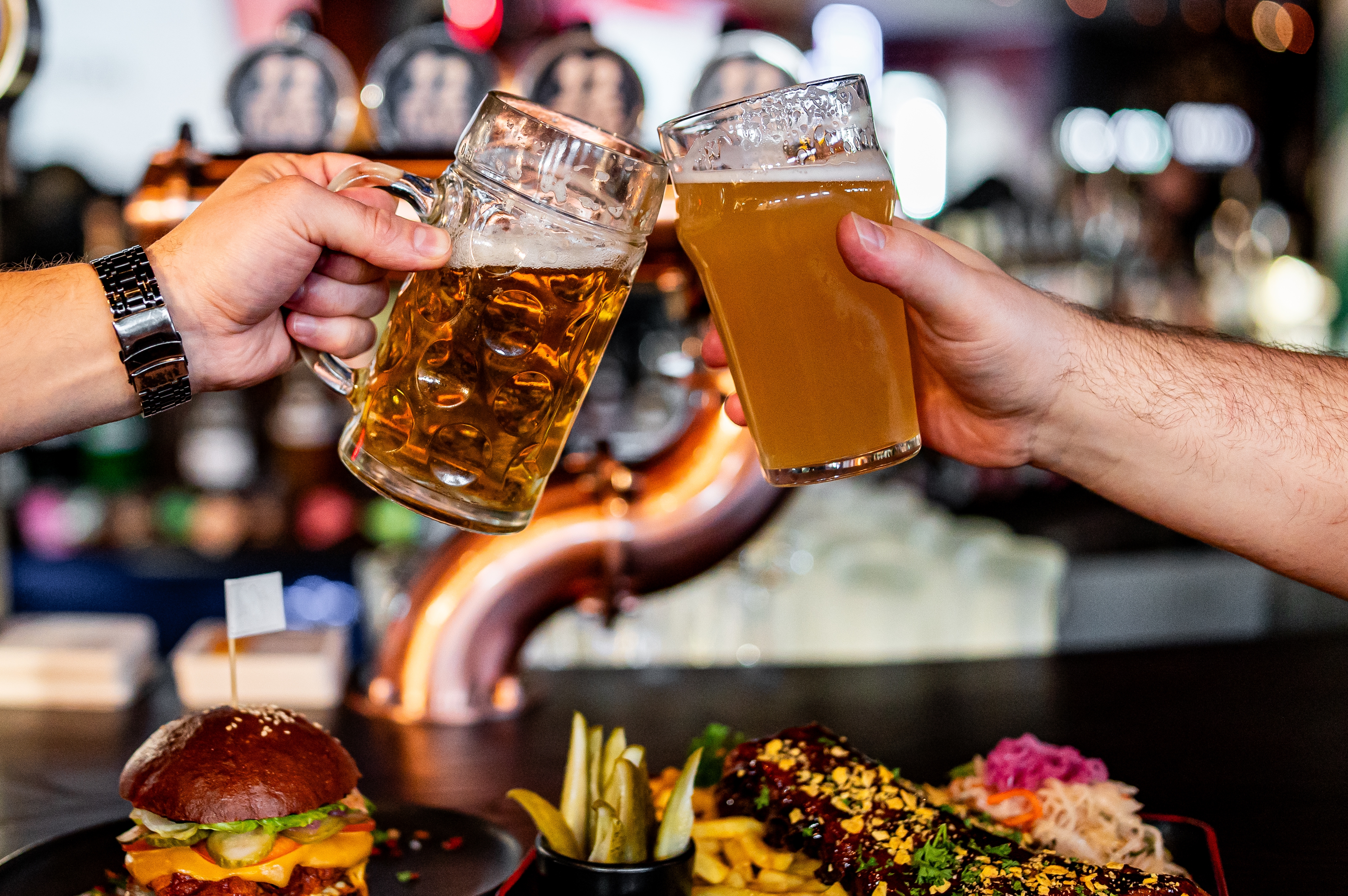 Two people clinking beer glasses over a table with a burger, fries, and a dish topped with herbs, suggesting a lively dining scene