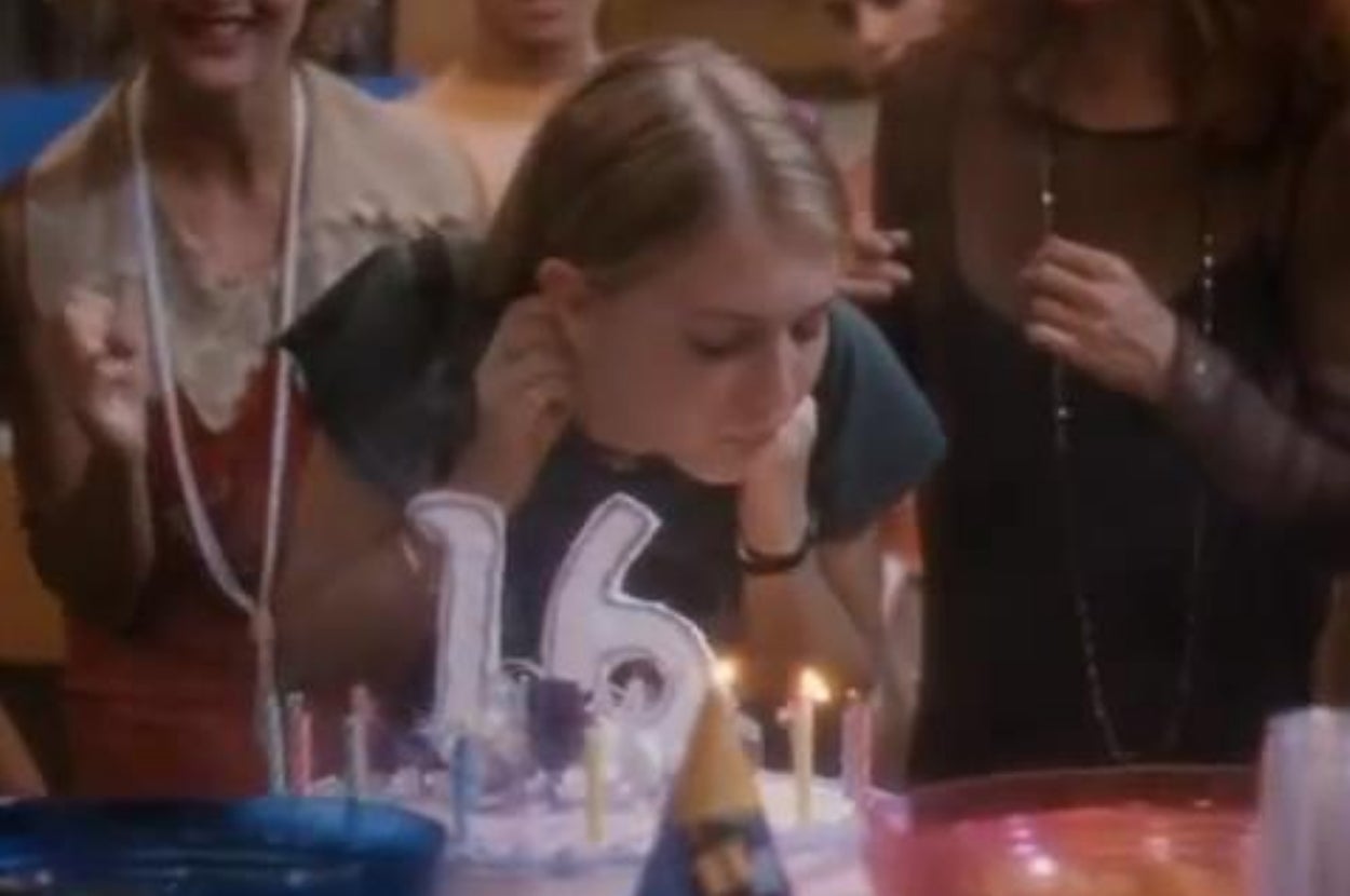 A young person surrounded by friends blows out candles on a birthday cake with the number 16