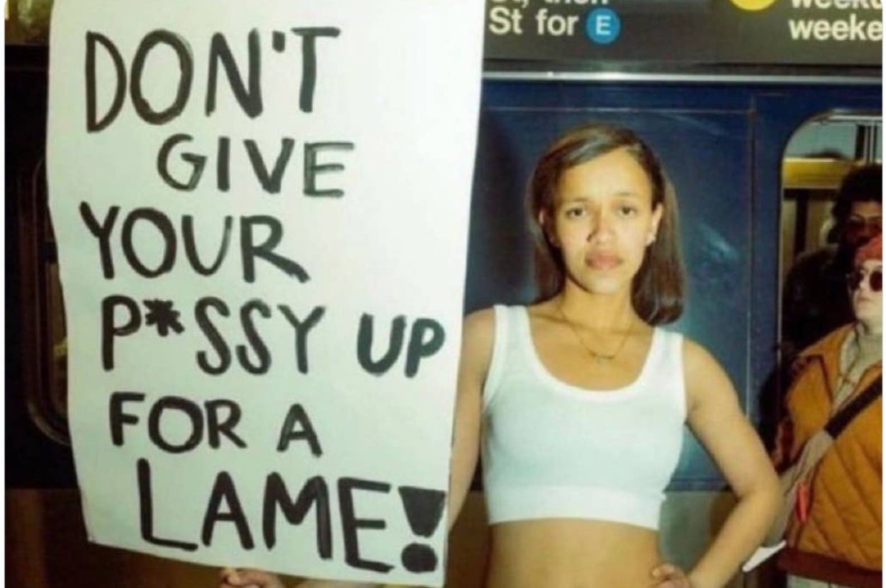 A woman stands in a subway holding a sign that reads, "DON'T GIVE YOUR P*SSY UP FOR A LAME!"
