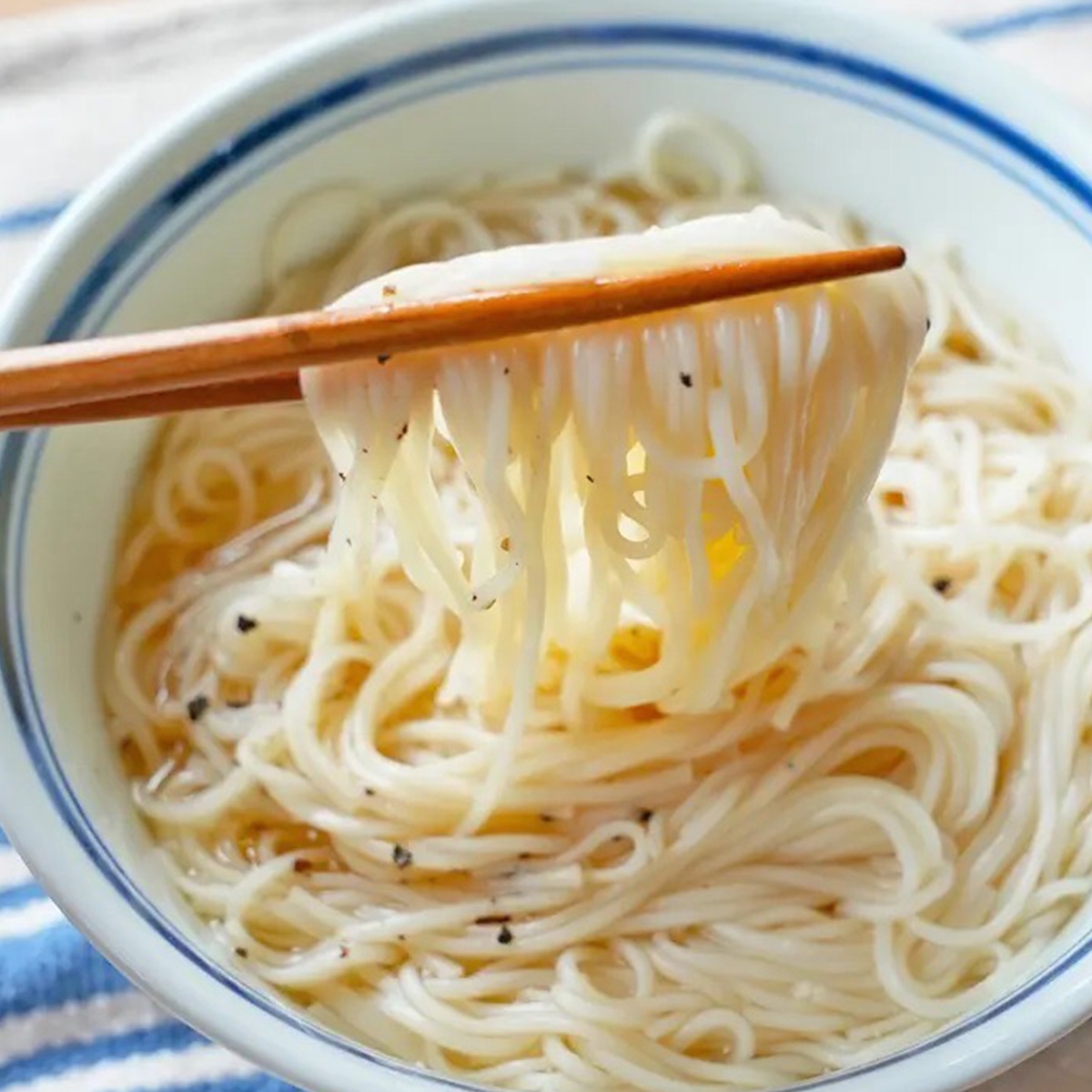 箸で持ち上げられた温かい麺の入った白いボウル。薄いスープに浸った素麺が見える。