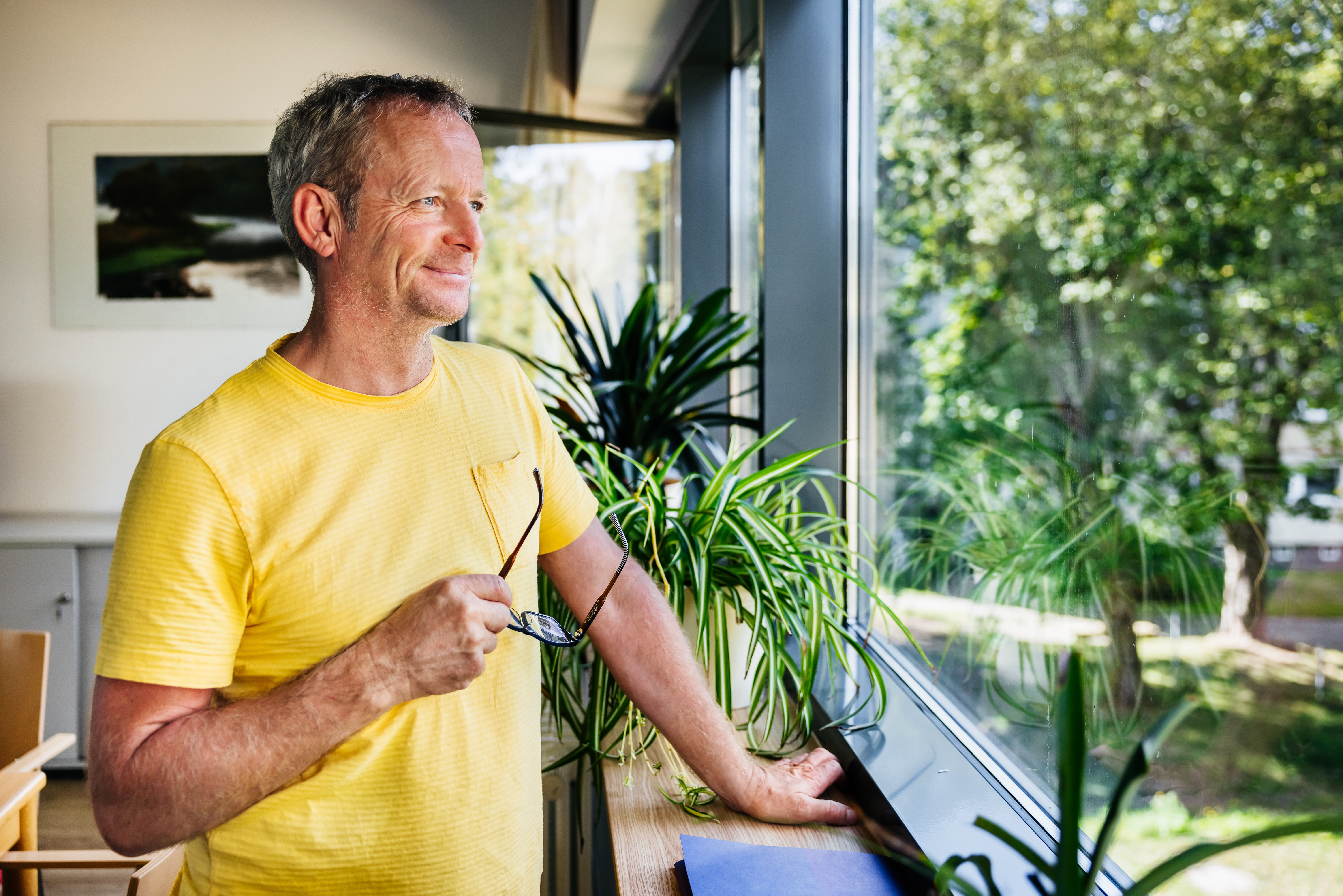 Mann i en uformell skjorte med kort ermet som holder briller, står ved et vindu med planter, stirrer ute ettertenksomt
