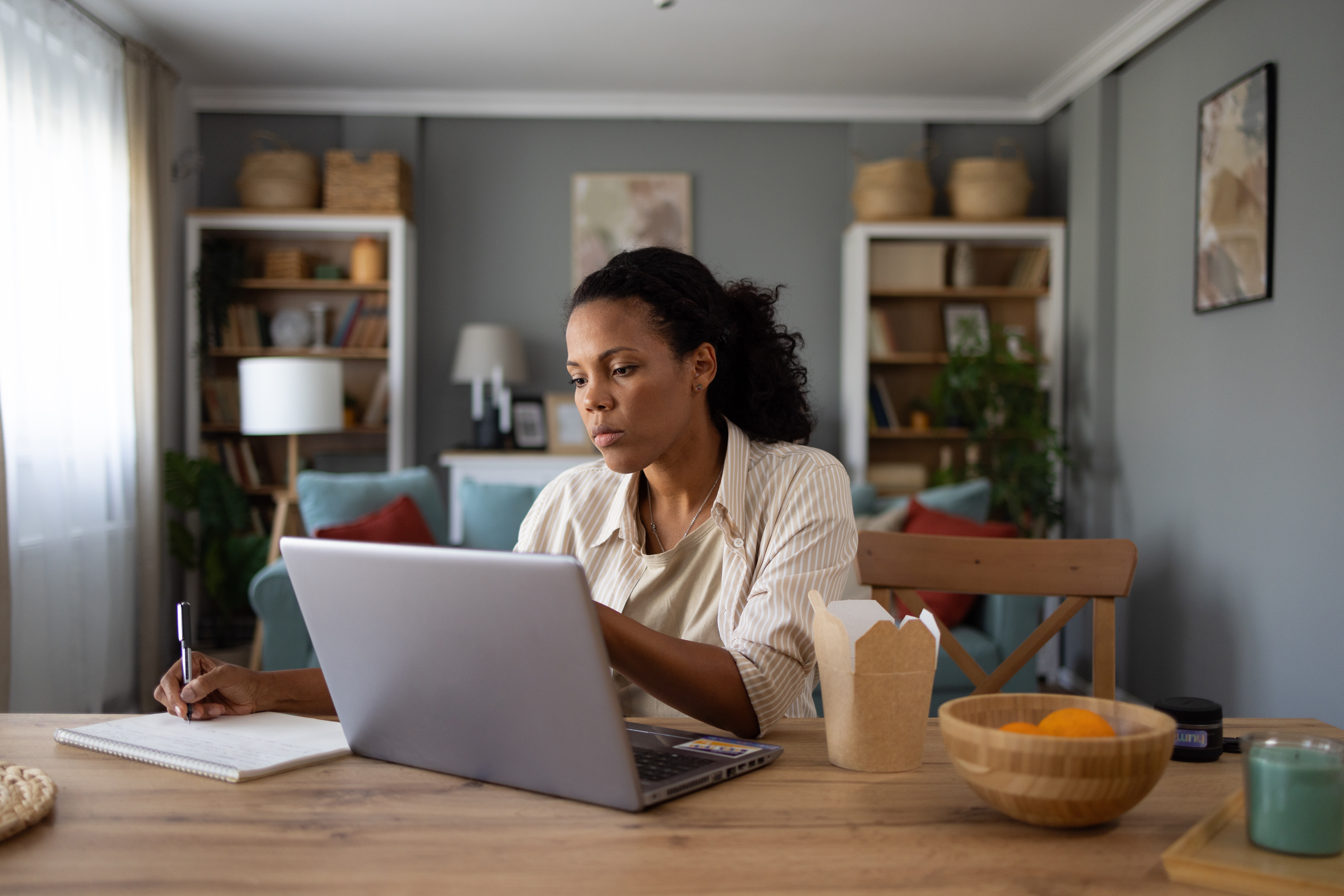 En person jobber på en bærbar datamaskin ved et spisebord med en åpen notisbok, takeaway -beholder og skål med appelsiner i nærheten, i en koselig stue