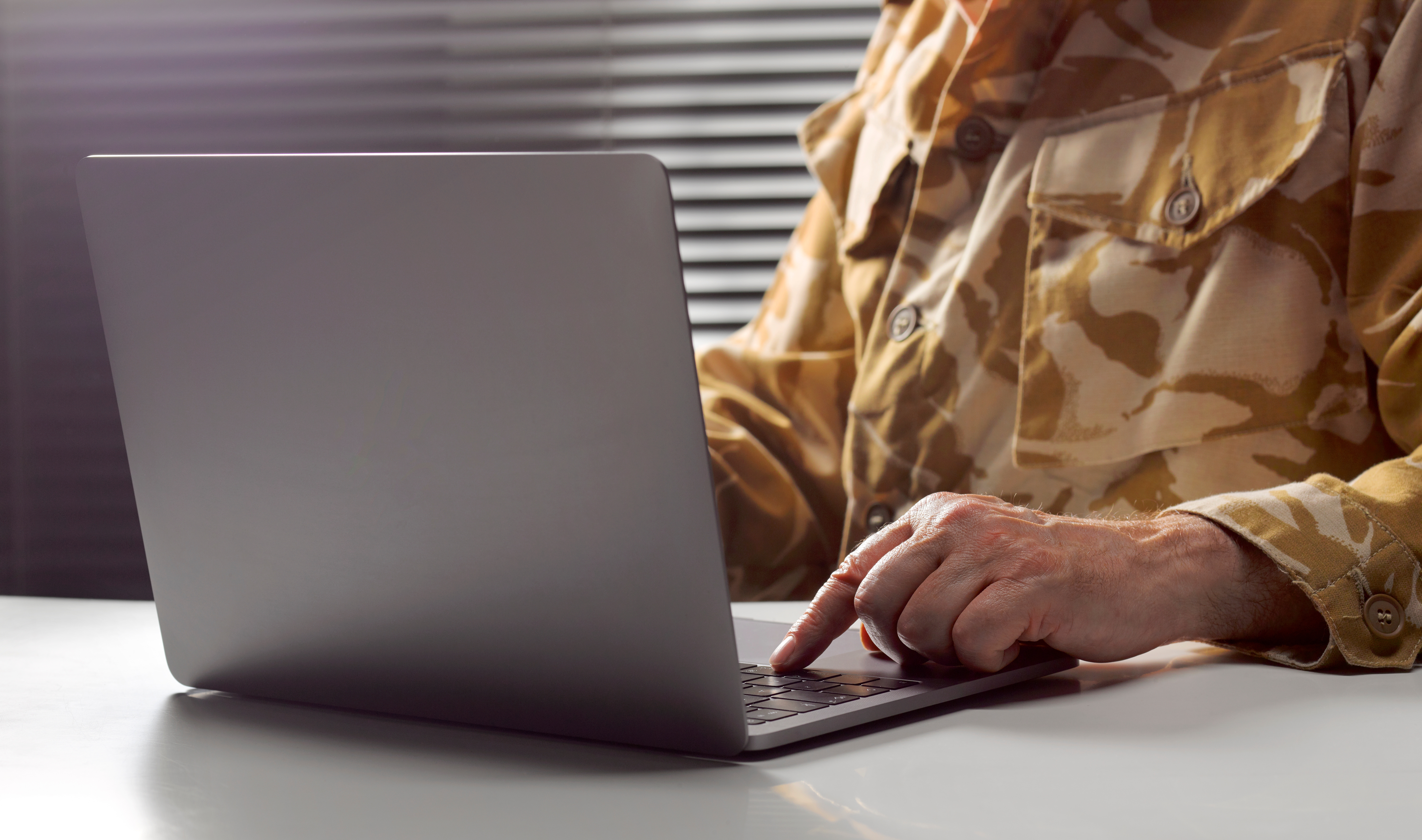 En person som har på seg en kamuflasjejakke på en bærbar datamaskin på et skrivebord, med persienner i bakgrunnen