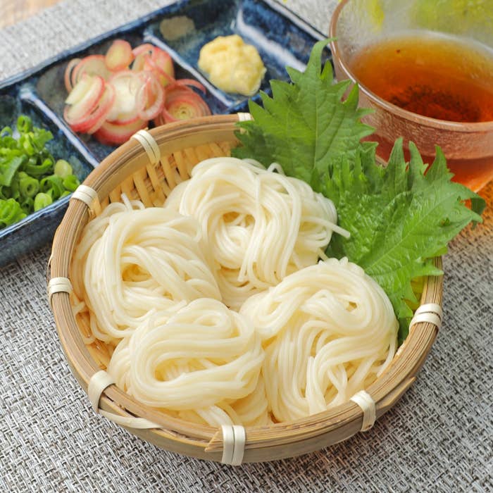 籠に盛られた素麺と薬味(ネギ、みょうが、生姜)と冷たい飲み物。夏の食卓をイメージさせる。