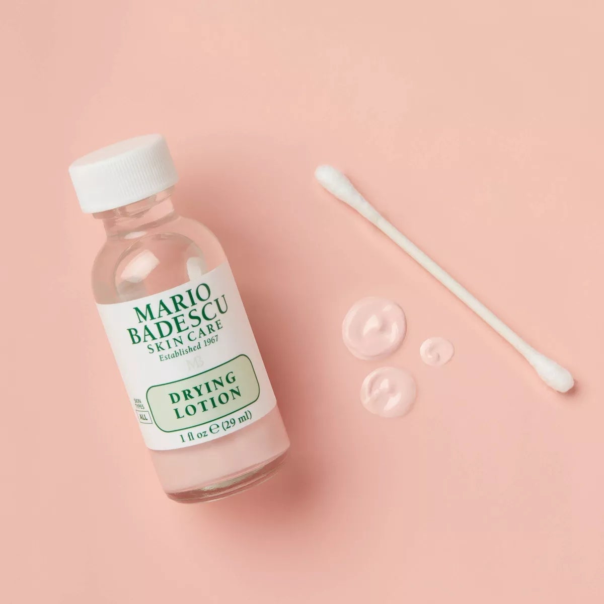 Botol lotion pengeringan Mario Badescu dengan tetesan produk di sebelah Q-Tip dengan latar belakang merah muda