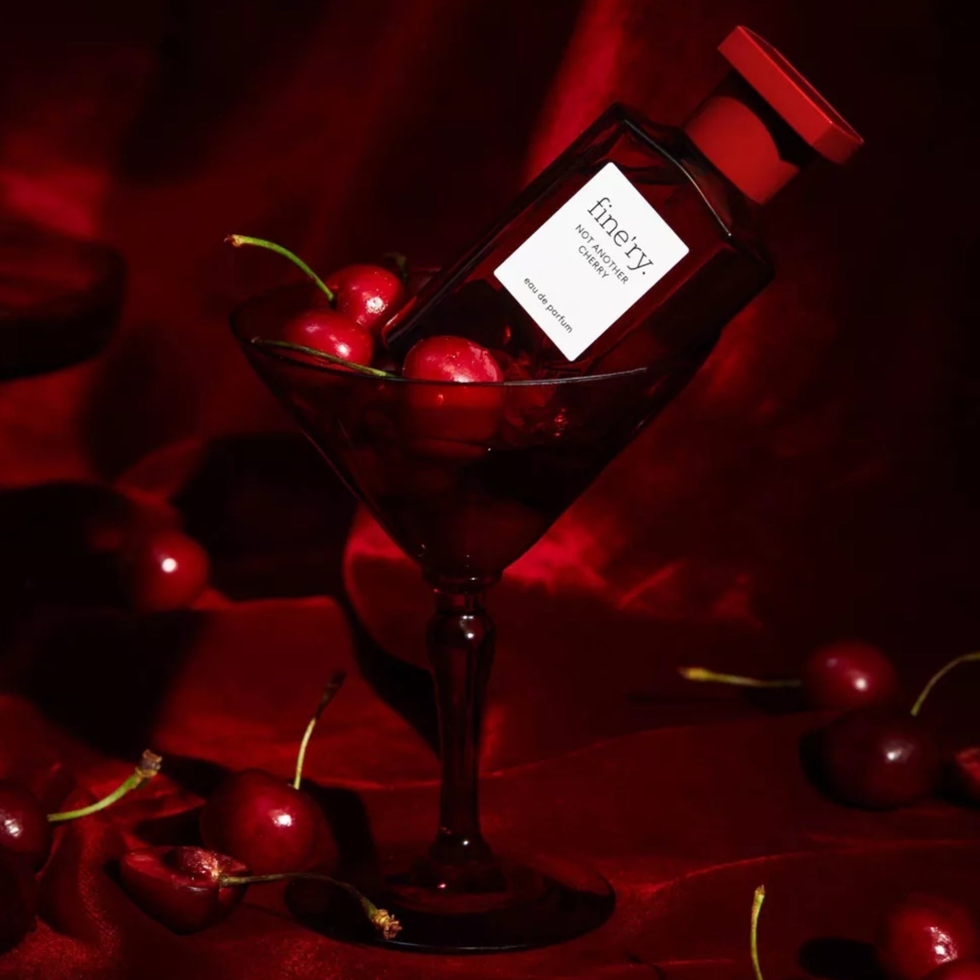 Botol parfum halus na martini glass dikelilingi oleh ceri, diletakkan dengan latar belakang merah anggur yang kaya