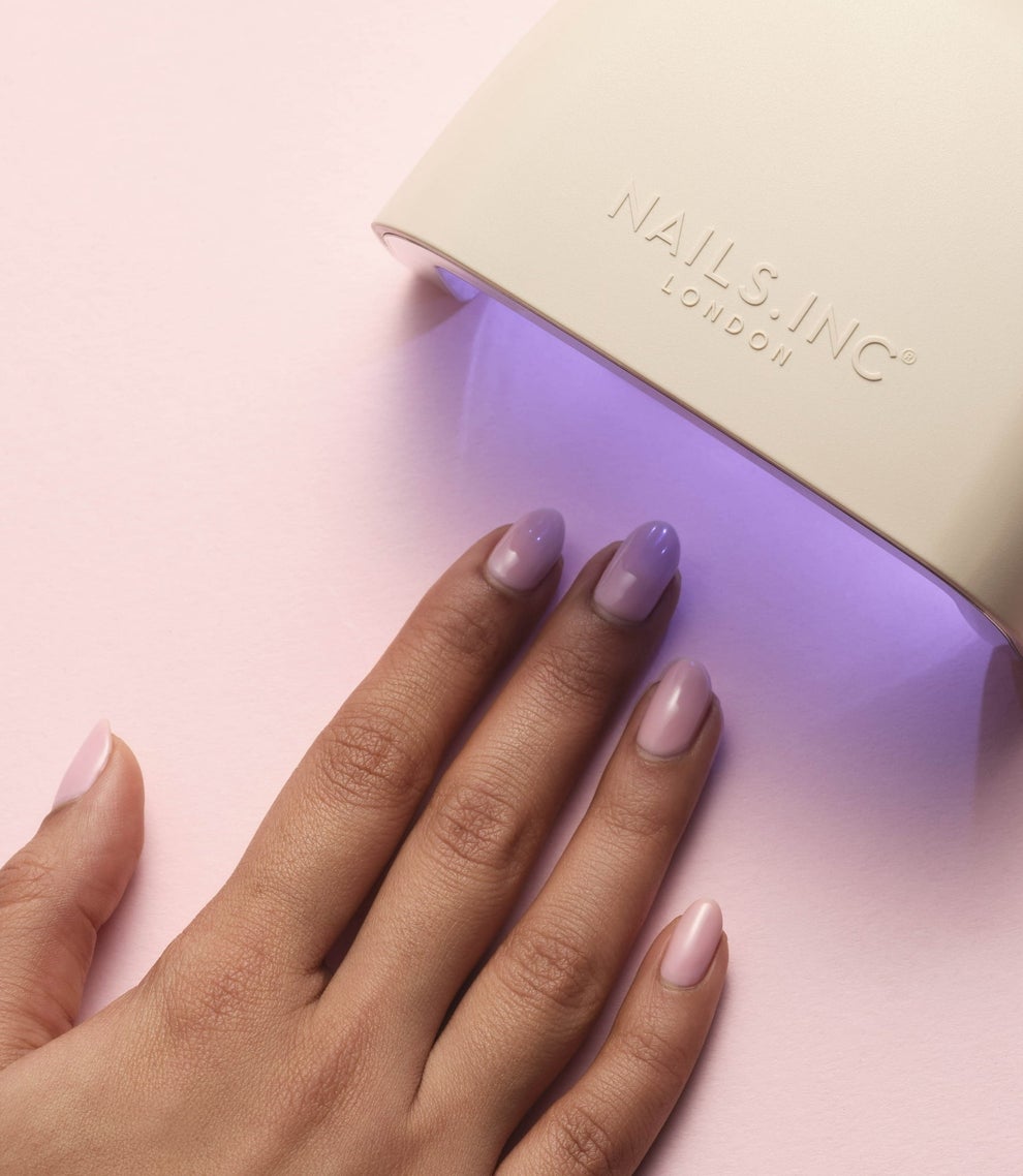 Hand with pastel nails under a UV lamp for drying; the lamp is branded with "NAILS INC London."