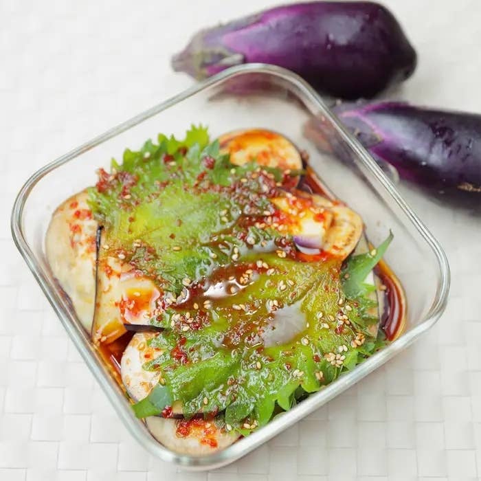 皿に盛られたなすの漬物、しそと調味料が添えられています。上品な盛り付けが印象的です。後ろには新鮮ななす。