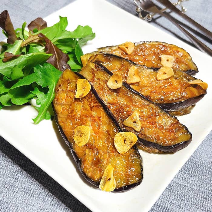 ナスのソテーにスライスガーリックをトッピングし、横に葉野菜が添えられた料理の写真。