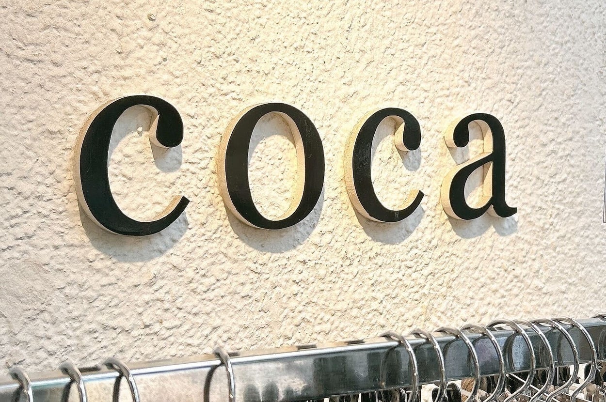 ショップの壁に「coca」と書かれたサインがあり、衣類がラックに並んでいる。