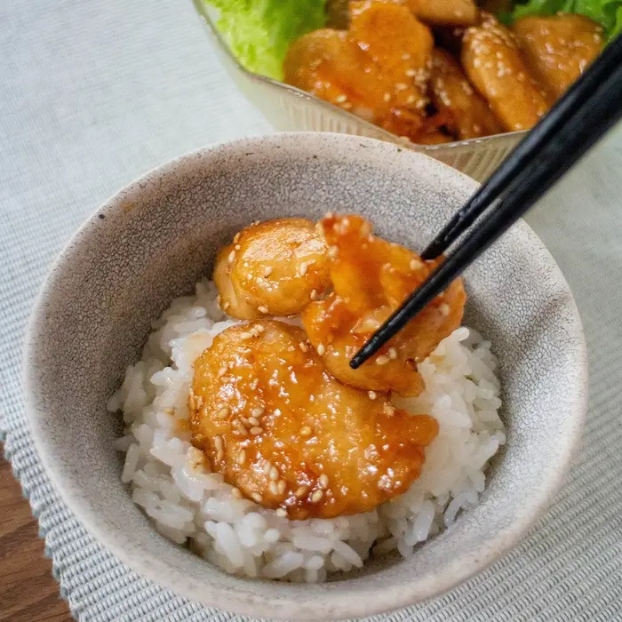 白いご飯の上に、箸で持ち上げた照り焼きチキンの様子。背景に似た料理の一皿が見える。